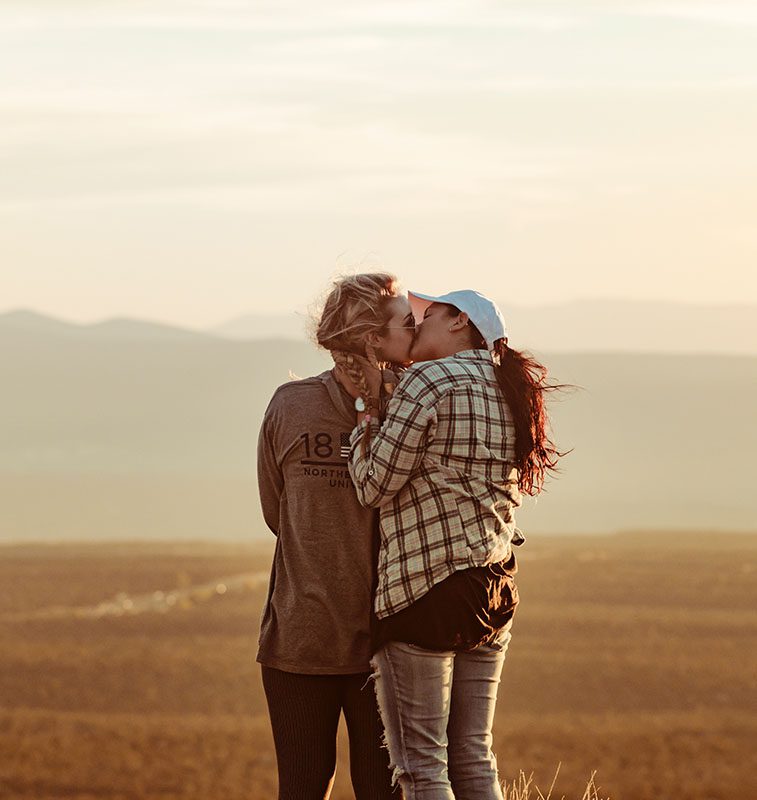 Two women kissing in a field with mountains behind them.