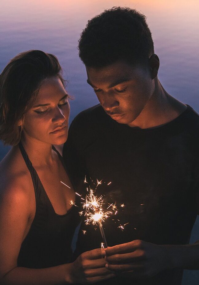 A man and woman holding sparklers in front of the ocean.